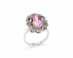 Ring with a pink oval center stone surrounded by a halo of clear stones and small green gems on a silver band