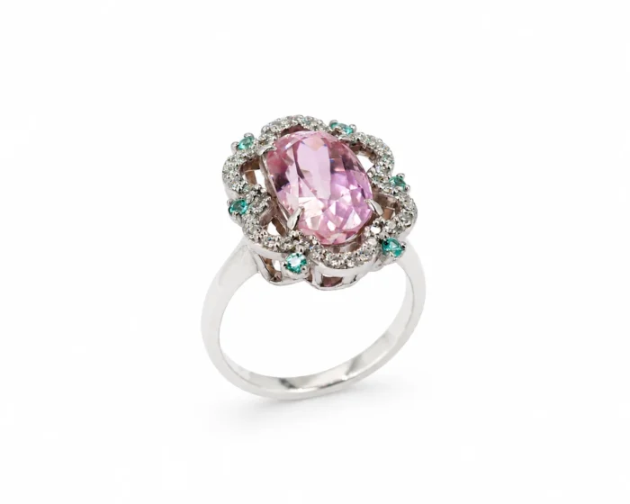 Ring with a pink oval center stone surrounded by a halo of clear stones and small green gems on a silver band
