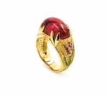 Gold ring with a large red oval gemstone, framed by decorative leaf-shaped gold settings and green/purple accent stones on the band.