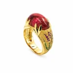 Gold ring with a large red oval gemstone, framed by decorative leaf-shaped gold settings and green/purple accent stones on the band.