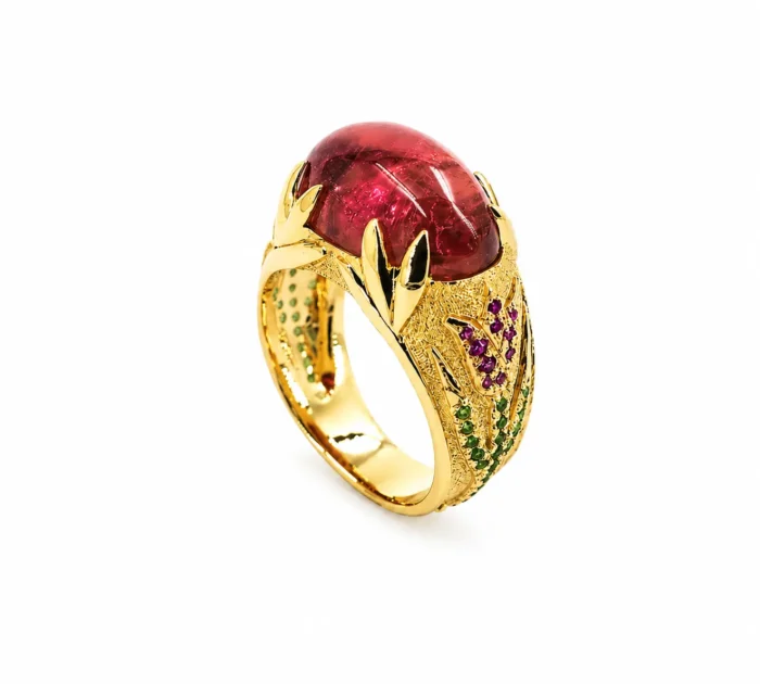 Gold ring with a large red oval gemstone, framed by decorative leaf-shaped gold settings and green/purple accent stones on the band.